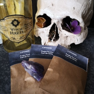 May include: Three brown paper bags with black labels that read "L'ATHANOR Poudre Hoodoo Can't Go Away" and a purple amethyst crystal on top of the bags. The bags are in front of a white human skull with a dried flower in each eye socket.