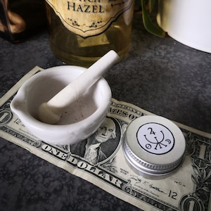 May include: A white ceramic mortar and pestle with a small amount of brown powder inside. The mortar and pestle are sitting on a one dollar bill with the text "ONE DOLLAR" visible. A small silver container with a black and white symbol is also on the bill.