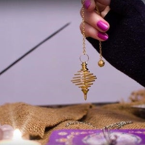 May include: A gold pendulum with a spiral design hangs from a chain held by a hand with pink nail polish. The pendulum is over a purple tarot card spread on a burlap surface. There is a lit candle and a small bag in the background.