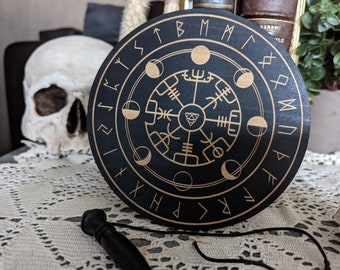 May include: A circular black wooden board featuring gold-colored engravings of the Vegvisir symbol at the center, surrounded by a ring of lunar phases and an outer border of runic characters. A black pendulum with a cord is positioned in the foreground.