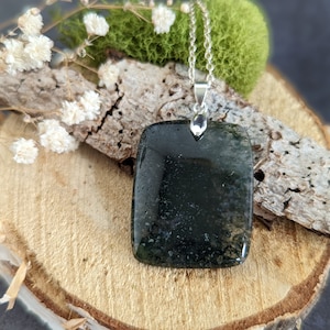 Puede incluir: Un colgante cuadrado de color verde oscuro y negro con una cadena de plata. El colgante está hecho de piedra natural y tiene una textura única y orgánica.