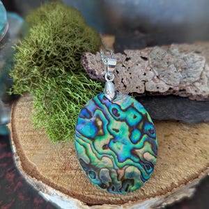Op de afbeelding: Ovale abalone schelpen hanger met een zilveren ketting. De hanger toont iriserende blauwe, groene en paarse kleuren. De ketting wordt gepresenteerd op een houten oppervlak met groen mos en een stuk steen op de achtergrond.