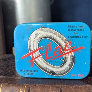May include: A vintage blue metal tin with the word "Fil'Lac" in red script, featuring a tire inner tube graphic. Text includes "Vulcanisation a Froid" and "Réparation instantanée des chambres à air."