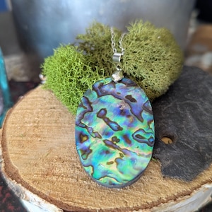 Peut inclure: Collier pendentif ovale avec coquillage d'ormeau irisé, aux nuances de bleu, vert et violet. Le pendentif est suspendu à une fine chaîne argentée. Le collier est présenté sur une tranche de bois avec de la mousse verte.