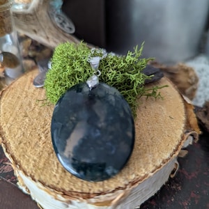 Peut inclure: Un collier en chaîne argent avec un grand pendentif ovale en agate mousse verte. Le pendentif est de couleur vert foncé avec des inclusions brunes et blanches. Le collier est présenté sur une surface en bois avec de la mousse verte.