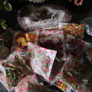 Könnte beinhalten: Sortiment getrockneter Kräuter und Blumen, verpackt in kleinen durchsichtigen Beuteln mit Blumenmustern. Die Beutel sind auf einer dunklen Oberfläche angeordnet.