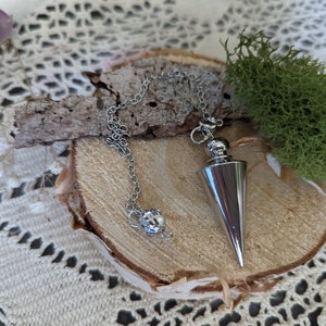May include: A silver metal pendulum with a pointed tip and a chain. The pendulum is hanging from a wooden surface with green moss.