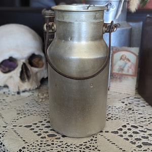 May include: A silver aluminium milk can with a rusty handle. The can has a slightly dented top and a smooth, shiny surface.