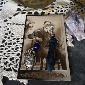 May include: A silver pendulum with a blue gemstone and a scarab beetle charm hangs over a vintage photograph. The pendulum is connected to a chain with a blue lapis lazuli bead. A purple crystal sits nearby.