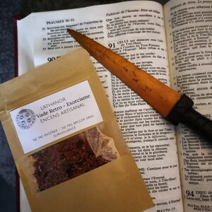 May include: A small brown paper bag with a label that reads "L'ATHANOR Vade Retro - Exorcism ENCENS ARTISANAL" and a warning to "NE PAS ING&Eacute;RER - NE PAS BR&Uuml;LER SANS SURVEILLANCE". The bag contains a red and brown mixture of incense. A wooden knife with a black handle is lying on top of the bag.