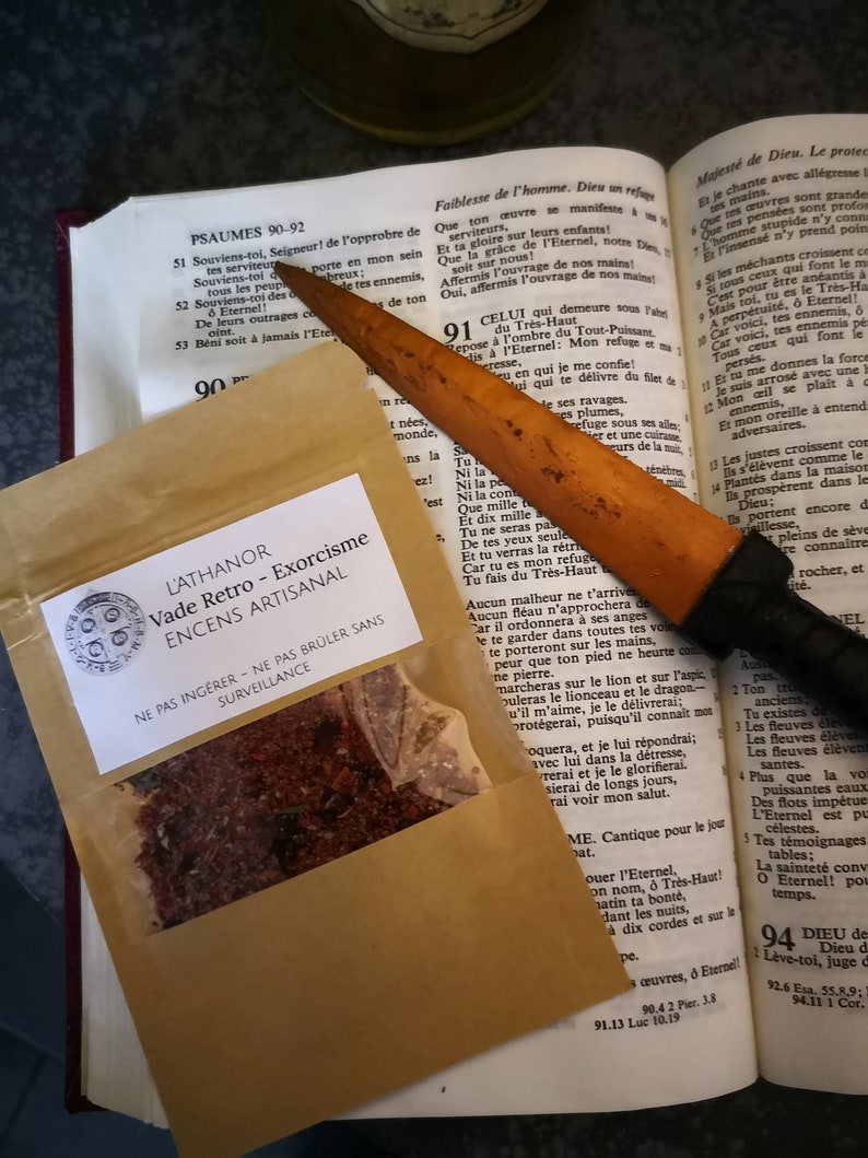 May include: A small brown paper bag with a label that reads "L'ATHANOR Vade Retro - Exorcism ENCENS ARTISANAL" and a warning to "NE PAS ING&Eacute;RER - NE PAS BR&Uuml;LER SANS SURVEILLANCE". The bag contains a red and brown mixture of incense. A wooden knife with a black handle is lying on top of the bag.