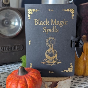 Peut inclure: Un livre noir intitulé "Black Magic Spells" avec des lettres et des illustrations dorées. Le livre présente un motif de bougie et de crâne. Un ruban noir est attaché. Le livre est présenté avec une petite citrouille orange et une lampe de poche vintage.