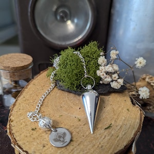 Puede incluir: Un péndulo de plata con una punta puntiaguda y una cadena con una bola decorativa. El péndulo está apoyado sobre una superficie de madera con musgo y flores blancas.