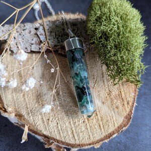 May include: A green and black crystal pendant on a silver chain. The pendant is shaped like a long, thin cylinder and is set on a wooden slice with moss.
