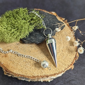 Puede incluir: Un péndulo de plata con una punta puntiaguda cuelga de una cadena de plata sobre una superficie de madera. El péndulo está rodeado de musgo verde y flores blancas.