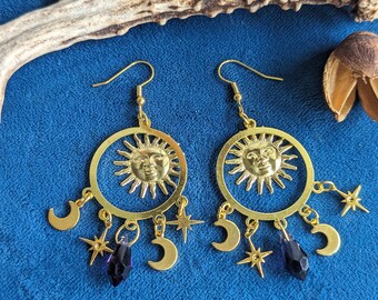 May include: Gold tone earrings with a sun and moon design. Each earring features a circular sun with a face, surrounded by smaller moons and stars. The earrings are accented with dark purple crystal drops.