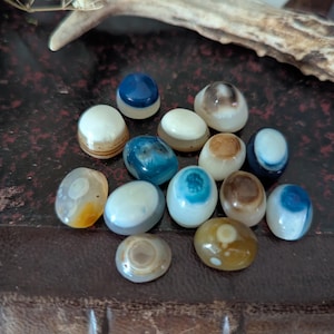 May include: A selection of polished, oval stones in shades of blue, brown, white, and yellow. The stones display unique patterns and are arranged on a dark, textured surface, possibly leather.