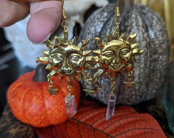 May include: Gold sun earrings with crystal accents. Each earring features a detailed sun face with a smiling expression and a dangling crystal charm. The earrings are suspended from gold hooks.