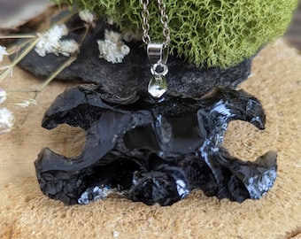 May include: A silver chain necklace with a black obsidian pendant. The pendant is a dark, irregularly shaped stone with a polished surface. The necklace is displayed on a wooden surface with green moss and small white flowers in the background.