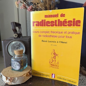 May include: A yellow book titled "manuel de radiesthésie" with red text, next to a small glass jar with a cork stopper and a vintage metal lantern. The book's cover features text and an illustration of a person. The jar sits on a wooden slice.