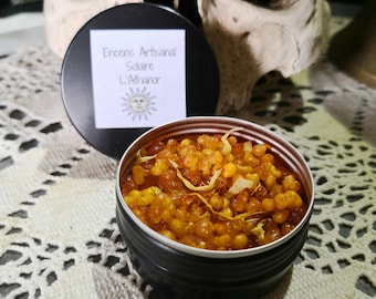 May include: A small, round, black tin container filled with orange and yellow resin incense. The lid of the tin is open and shows a label that reads "Encens Artisanal Solaire L'Athanor" with a sun graphic.