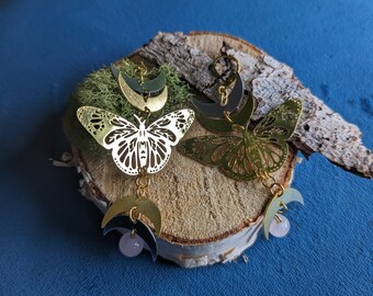May include: Gold butterfly earrings with a crescent moon design and a pink stone accent. The earrings are hanging from a wooden surface.
