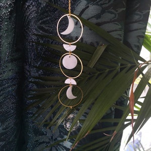 May include: A gold metal moon phase mobile with white ceramic moon and crescent shapes hanging from a chain. The mobile is hanging in front of a dark green patterned curtain.