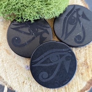Könnte beinhalten: Drei schwarze Untersetzer mit einem erhabenen Design des Horusauges. Die Untersetzer sind rund und haben eine glatte, polierte Oberfläche.
