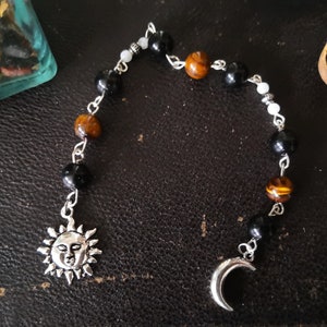 Könnte beinhalten: Eine silberne Kette mit schwarzen, braunen und weißen Perlen, einem Sonnenanhänger und einem Halbmondanhänger.