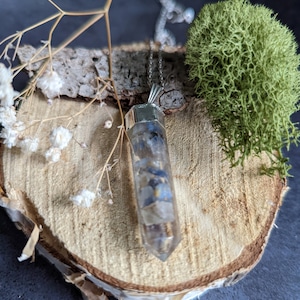 May include: A silver pendant necklace with a clear quartz crystal point that has blue and white inclusions. The pendant is on a wooden surface with moss and dried flowers.