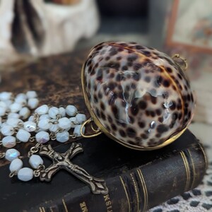 May include: A white rosary necklace with a silver cross charm rests on a brown leather-bound book. A decorative shell box with a brown and black spotted pattern sits on top of the book.
