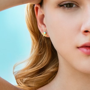 Op de afbeelding: Een close-up van het gezicht van een vrouw met een gouden, bloemvormige oorbel met een turquoise steen in het midden.