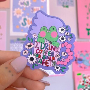 May include: A vibrant sticker with a green frog holding a knife and a sign saying "I DRINK COFFEE FOR YOUR SAFETY." The sticker has a purple border with flowers and a light purple background.