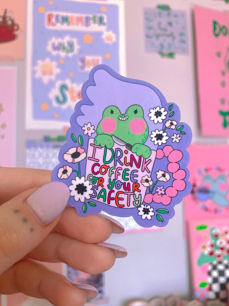 May include: A vibrant sticker with a green frog holding a knife and a sign saying "I DRINK COFFEE FOR YOUR SAFETY." The sticker has a purple border with flowers and a light purple background.