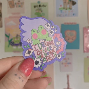 May include: A sticker featuring a green frog holding a coffee cup with the text "I drink coffee for your safety." The sticker has a purple border with white flowers and is held in a hand with red glitter nail polish.