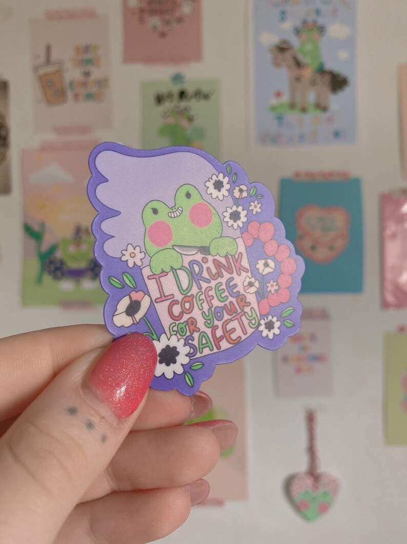 May include: A sticker featuring a green frog holding a coffee cup with the text "I drink coffee for your safety." The sticker has a purple border with white flowers and is held in a hand with red glitter nail polish.