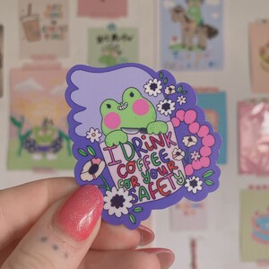 May include: A sticker with a green frog and pink cheeks holding a sign saying "I DRINK COFFEE FOR YOUR SAFETY." The sticker has a purple border with white flowers. Other decorative cards are in the background.