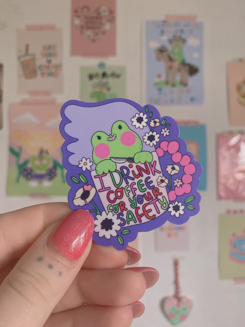 May include: A sticker with a green frog and pink cheeks holding a sign saying "I DRINK COFFEE FOR YOUR SAFETY." The sticker has a purple border with white flowers. Other decorative cards are in the background.