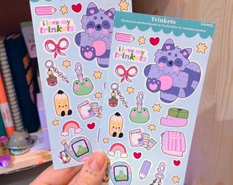 May include: Two sheets of stickers featuring a cartoon raccoon with the text "I love my trinkets". The stickers also include a frog, a pencil, a rainbow, a plant, a key chain, and a small handheld game.