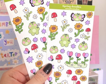 May include: Two sticker sheets with a spring theme. Each sheet features illustrations of a green frog, sunflowers, mushrooms, butterflies, flowers, and stars. The text "Spring doodles" and "Arteafoxstudios" is printed on the top of each sheet.