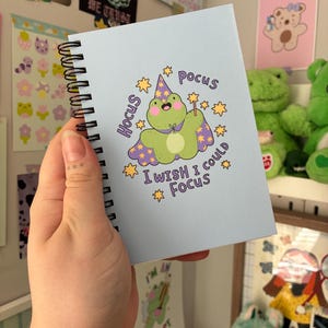 May include: A light blue spiral-bound notebook featuring a cartoon frog wearing a purple wizard hat and cape, holding a star-tipped wand. The text reads Hocus Pocus I WISH I could Focus, surrounded by yellow star illustrations on the front cover.