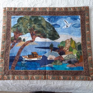 以下が含まれることがあります： 青、緑、茶色のカラーリングで構成された、額縁付きのキルティングされた壁掛け。シーンは、「Filou」と書かれた船、空を飛ぶ鳥、太い幹の木を描いています。