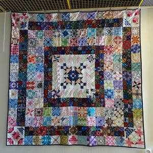 Könnte beinhalten: Eine große Decke aus vielen bunten Stoffquadraten mit verschiedenen Mustern und Designs. Die Decke ist im Patchwork-Stil gefertigt, wobei jedes Quadrat eine andere Farbe und ein anderes Muster hat. Die Decke ist von einem dunklen Rand umgeben.