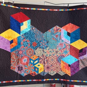 Może przedstawiać: Kolorowy koc z geometrycznym wzorem przedstawiającym wiele sześcianów wykonanych z różnych tkanin. Koc jest wykonany z różnych kolorów, w tym czerwonego, niebieskiego, żółtego, fioletowego i zielonego. Koc jest oprawiony w czarną ramkę i wielokolorową lamówkę.