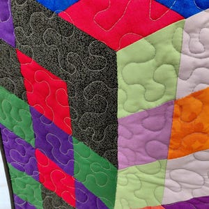 May include: Close-up of a colorful quilt with geometric shapes. The quilt features a variety of colors, including red, green, purple, and blue, with a black and white patterned fabric. The quilt is stitched with white thread in a wavy pattern.