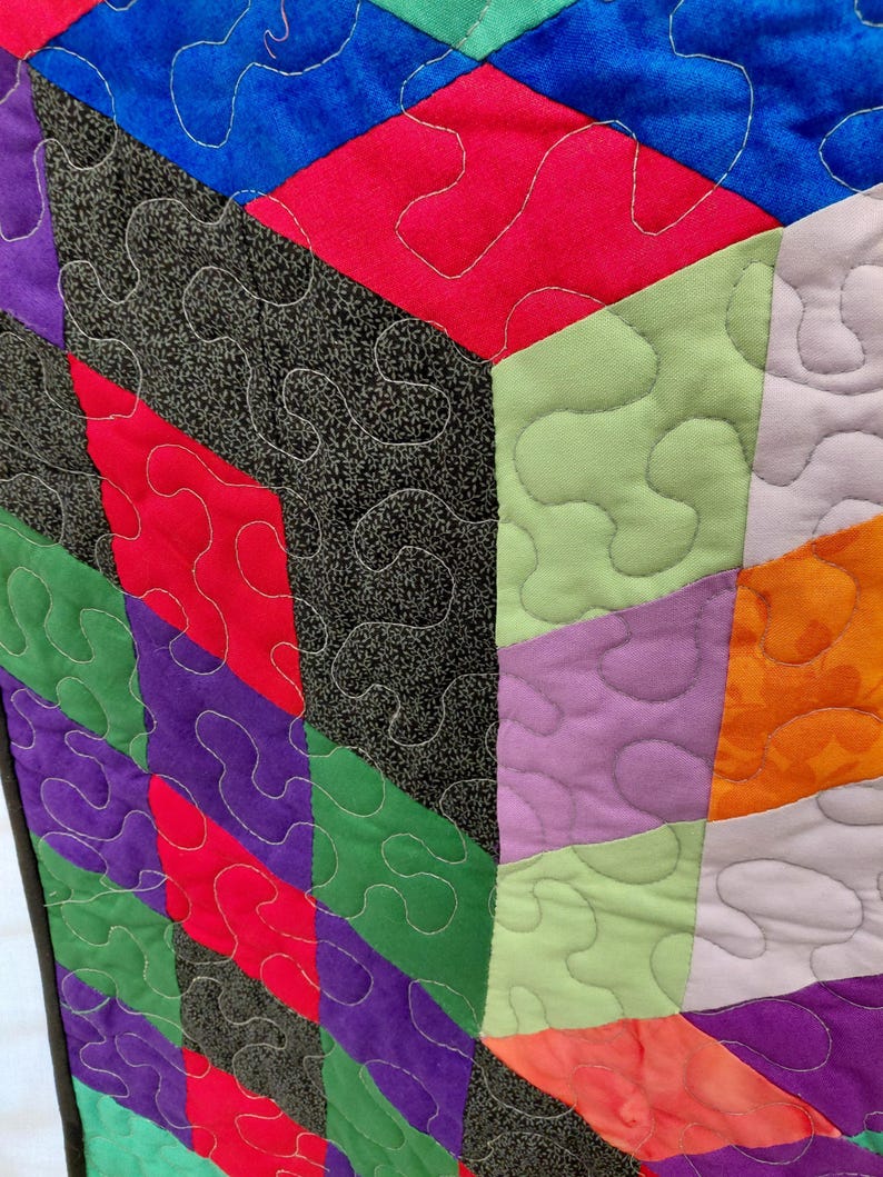 May include: Close-up of a colorful quilt with geometric shapes. The quilt features a variety of colors, including red, green, purple, and blue, with a black and white patterned fabric. The quilt is stitched with white thread in a wavy pattern.