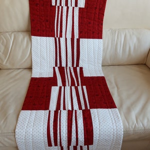 Puede incluir: Un camino de mesa acolchado rojo y blanco con un patrón geométrico. El camino de mesa está hecho de tela con un pequeño patrón de lunares blancos sobre un fondo rojo.