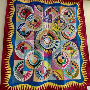 May include: A colorful quilt with a sunburst design. The quilt features a variety of geometric shapes and patterns in shades of red, blue, yellow, green, and pink. The quilt is framed with a red border and a yellow zigzag edge.