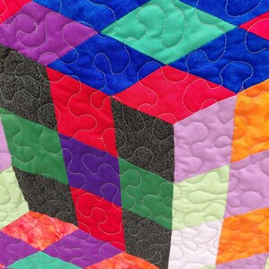 May include: Close-up of a colorful quilt with a geometric design. The quilt features diamond and rectangular shapes in shades of blue, green, red, purple, orange, and black. The quilt is machine quilted with white thread, creating a textured surface.