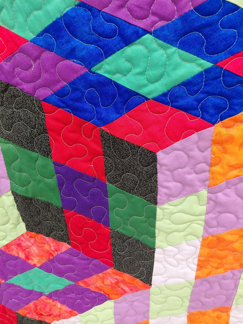 May include: Close-up of a colorful quilt with a geometric design. The quilt features diamond and rectangular shapes in shades of blue, green, red, purple, orange, and black. The quilt is machine quilted with white thread, creating a textured surface.